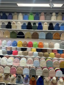 Wall display of stylish winter hats and beanies in various colors, designs, and materials, including pom-pom hats and knitted caps, showcasing diverse headwear styles.